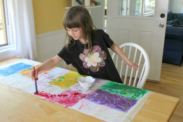 A young girl painting watercolors on a paper that has been scribbled on with oil pastels.