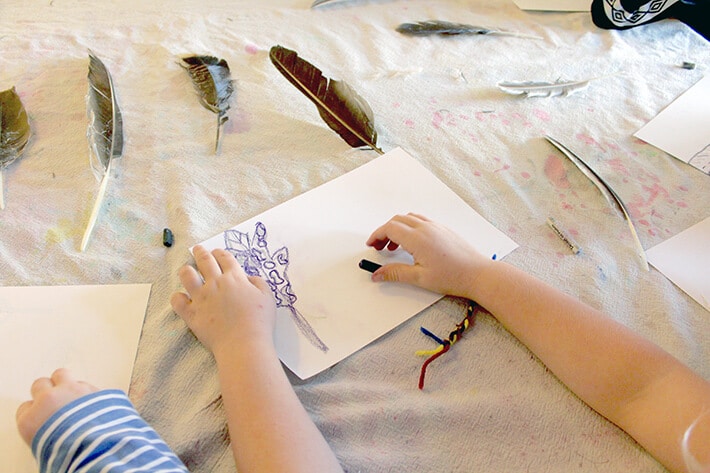 Student holding oil pastel drawing feather on paper with feathers laying on table in this easy nature drawing idea for kids