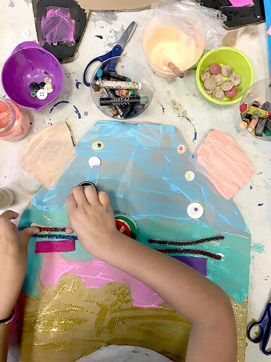 Child using paint, buttons, oil pastels, yarn, scissors and glue to create Miró inspired animal masks for kids