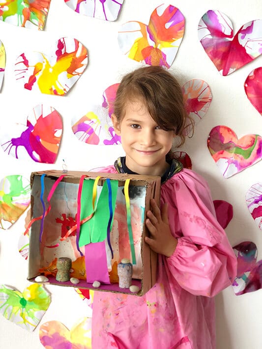 Girl holding shoebox diorama and standing in front of spin art heart wall