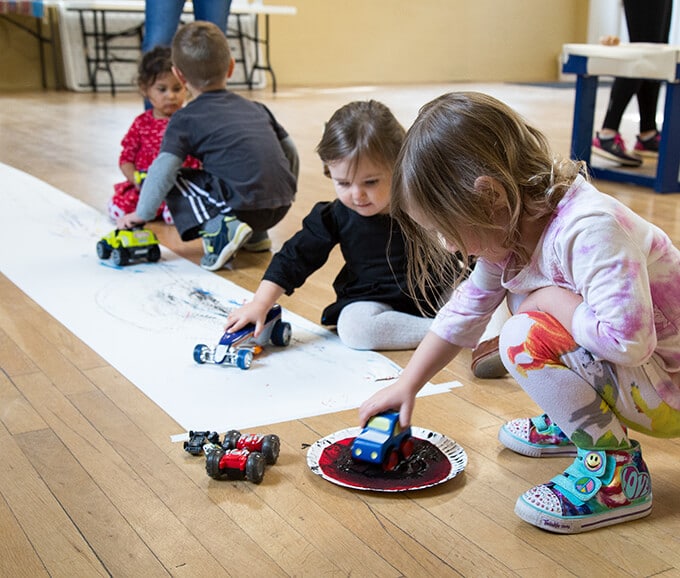 Kids roll cars in paint to create painted tracks on a roll of paper