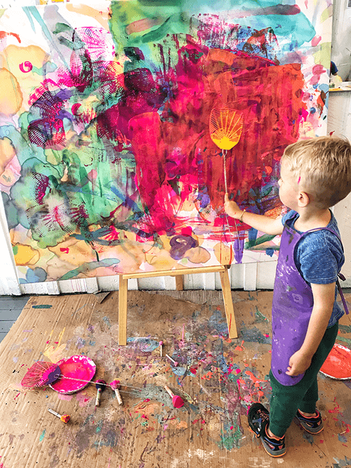 Child swatting paint onto canvas with fly swatter in kids art studio