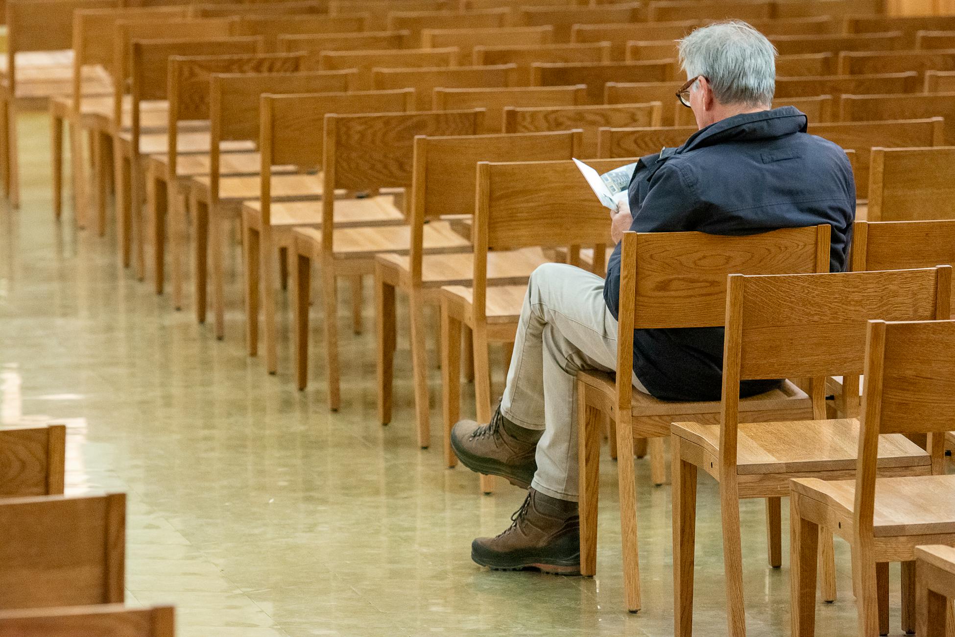 An elderly man sits reading a book in an empty church hall filled with wooden chairs.