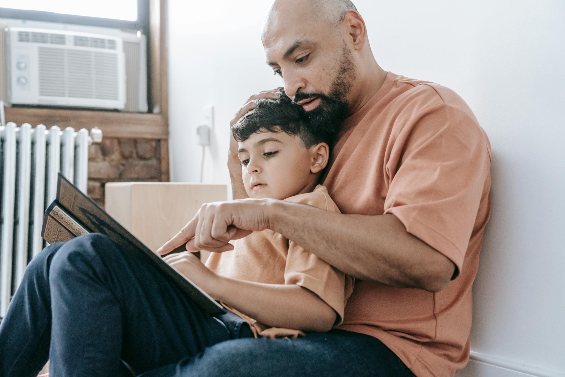 A father in an orange shirt reads a book to his son while sitting indoors, fostering learning and bonding.