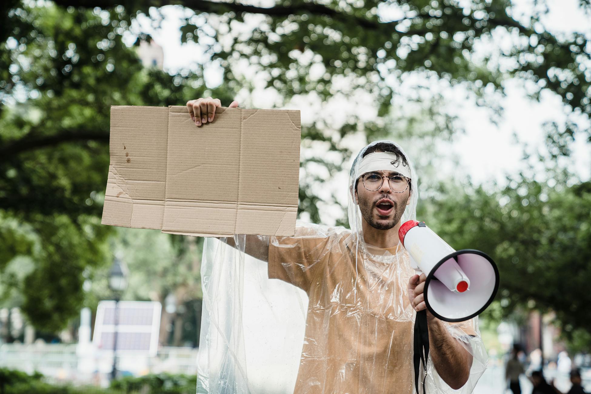 A passionate protestor wearing a rain poncho holds a blank sign and uses a megaphone outdoors.