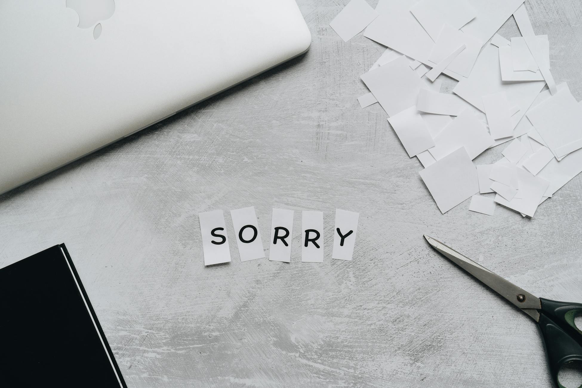 Top view of a desk with cut-out letters spelling 'sorry', scissors, and a laptop.