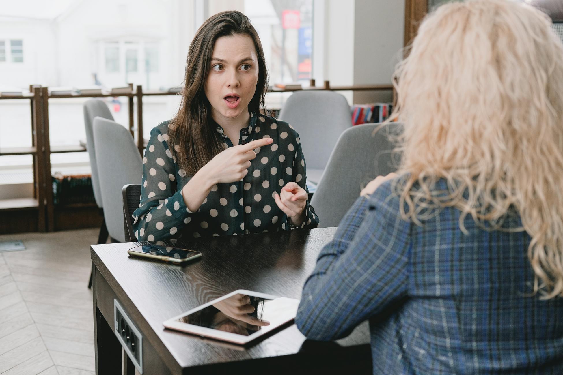 Two women in a lively discussion at a café, with expressive hand gestures and mobile devices on the table.