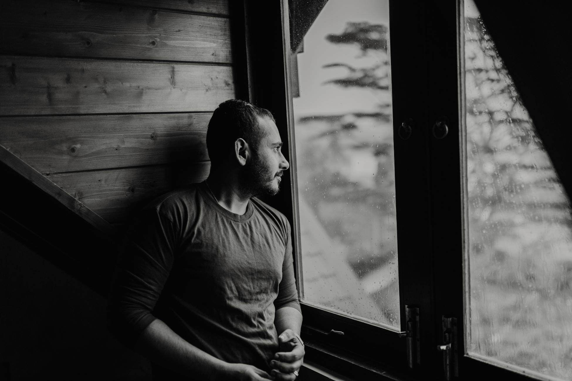 A bearded man thoughtfully gazes outside a window on a rainy day.