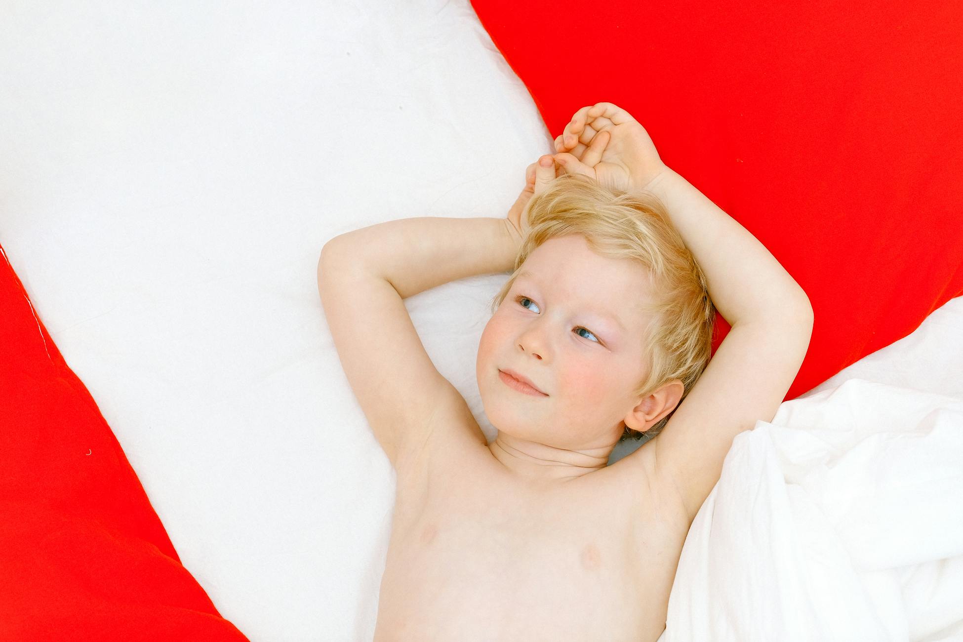 Adorable blond boy lying comfortably on a red and white bed, gazing thoughtfully.