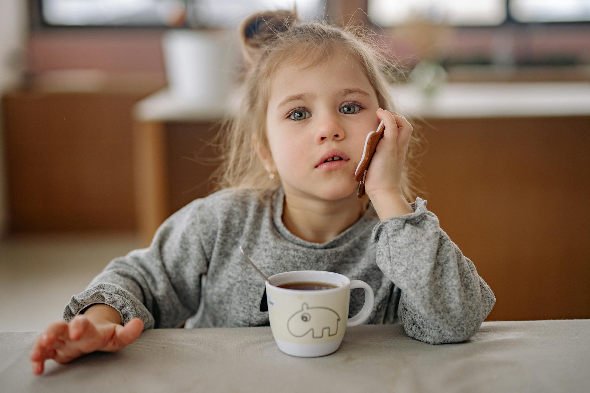 A young girl with blond hair sitting indoors, enjoying a drink with a thoughtful expression.