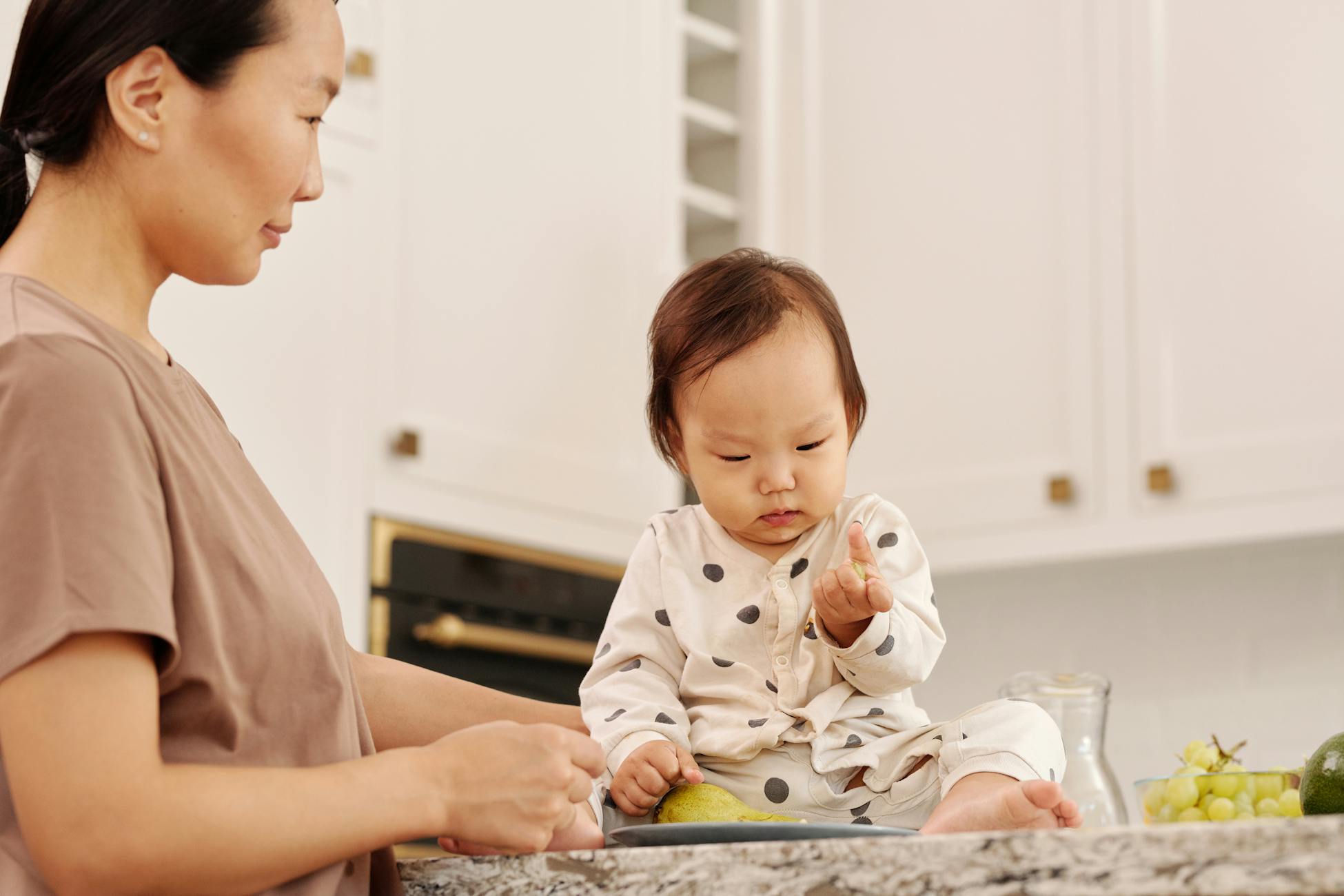 Asian mother and baby girl in a kitchen, sharing a tender moment. Bright and warm setting.