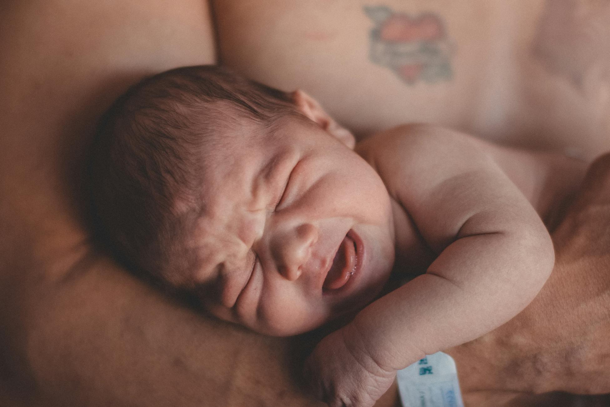 Tender moment of a newborn crying while cradled against a tattooed adult chest.