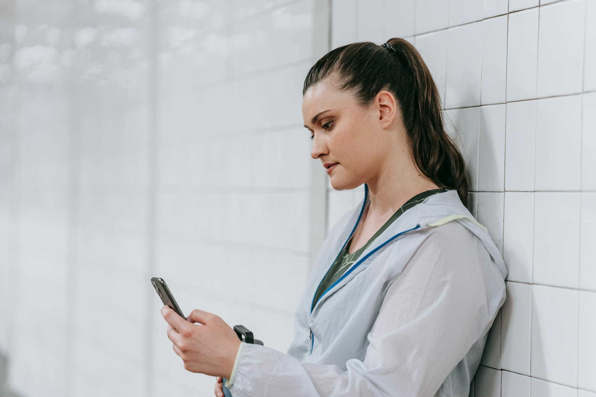 A young woman leans against a white wall while checking her smartphone indoors.