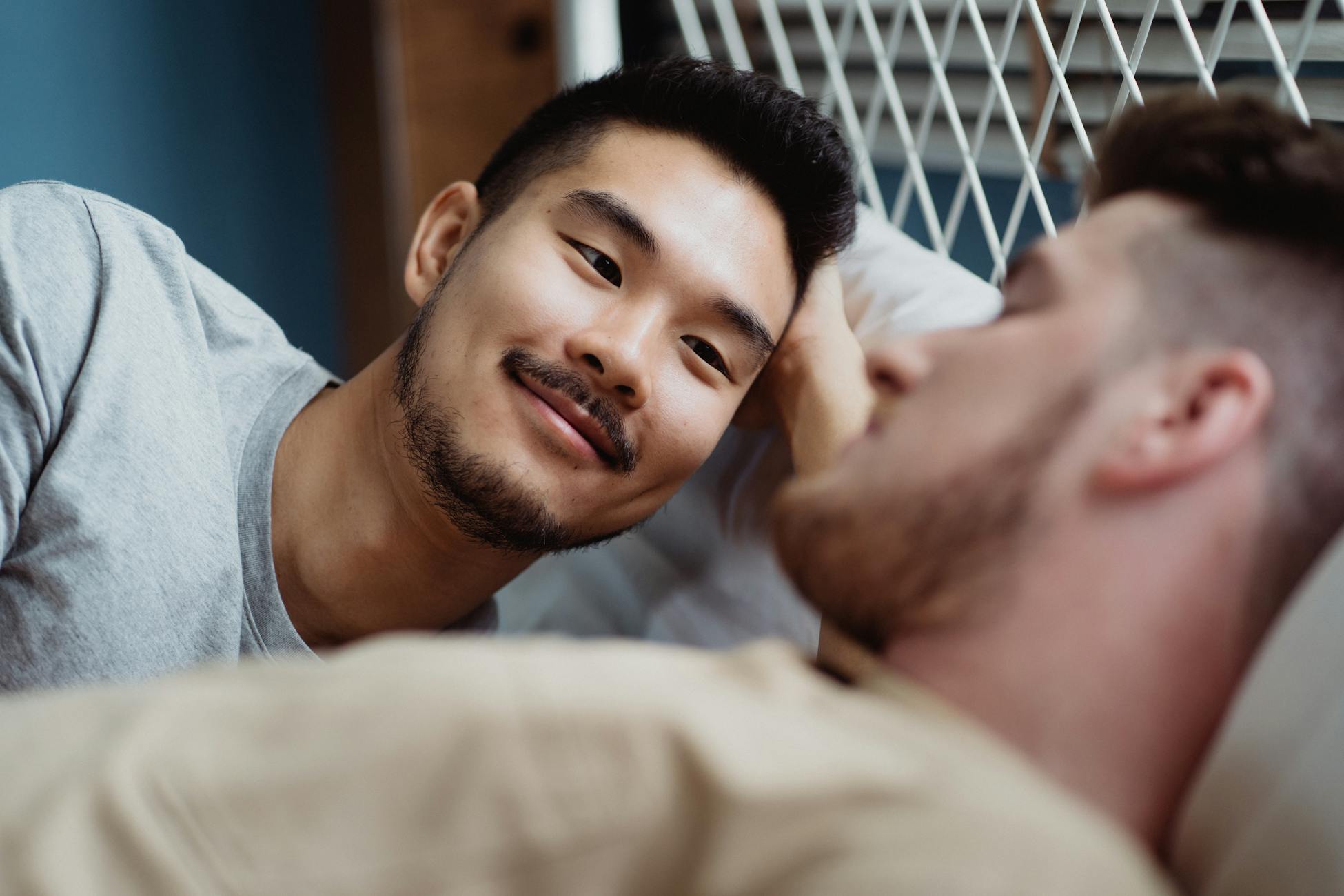 Close-up of an interracial gay couple sharing an intimate moment in bed, one partner awake and smiling.
