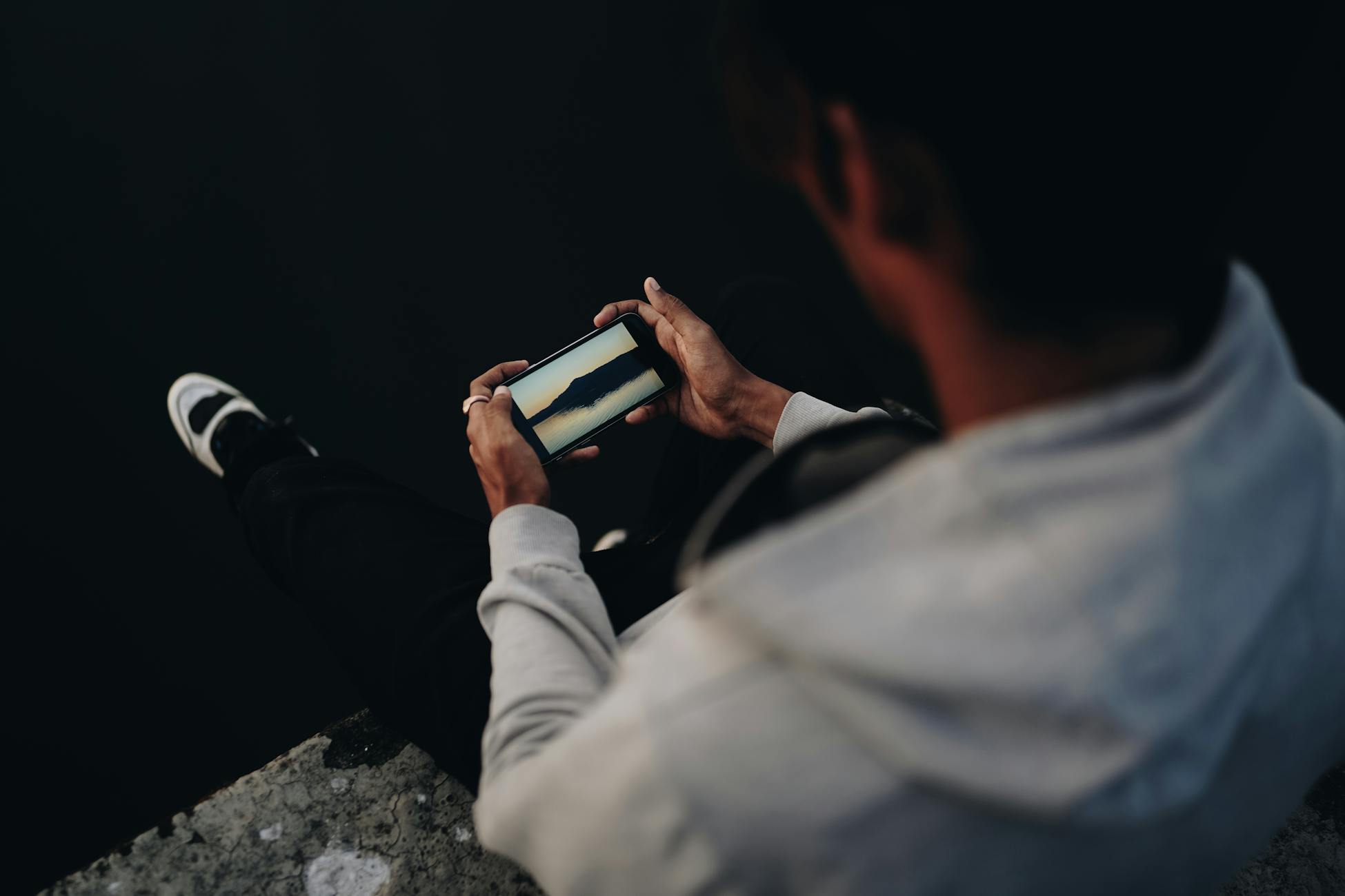 A man in casual attire sitting by the water using a smartphone, captured in a low-light setting.