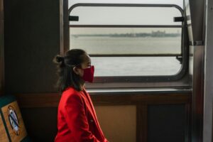 A woman in a red suit and face mask sits alone on a ferry, contemplating the view outside the window.