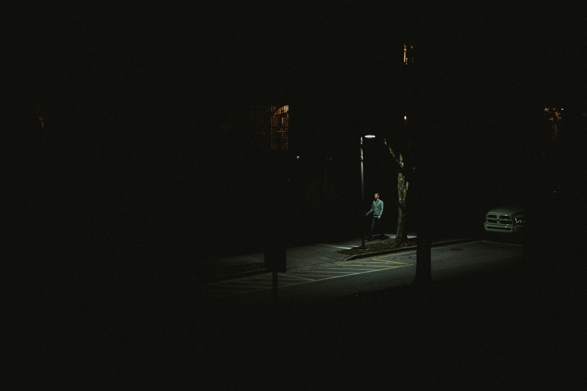 A lone person walking under a streetlight on a dark city street at night.