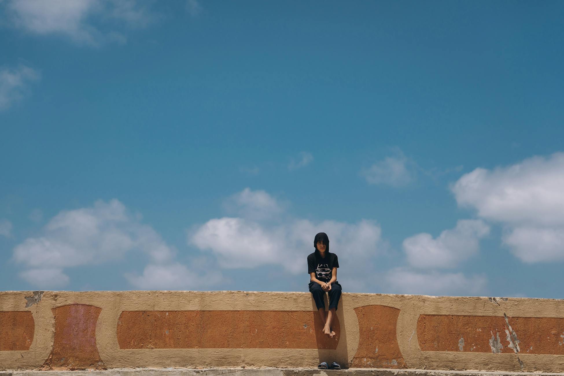 A teenager sits alone on a wall against a vibrant blue sky, capturing a moment of solitude and freedom in Egypt.