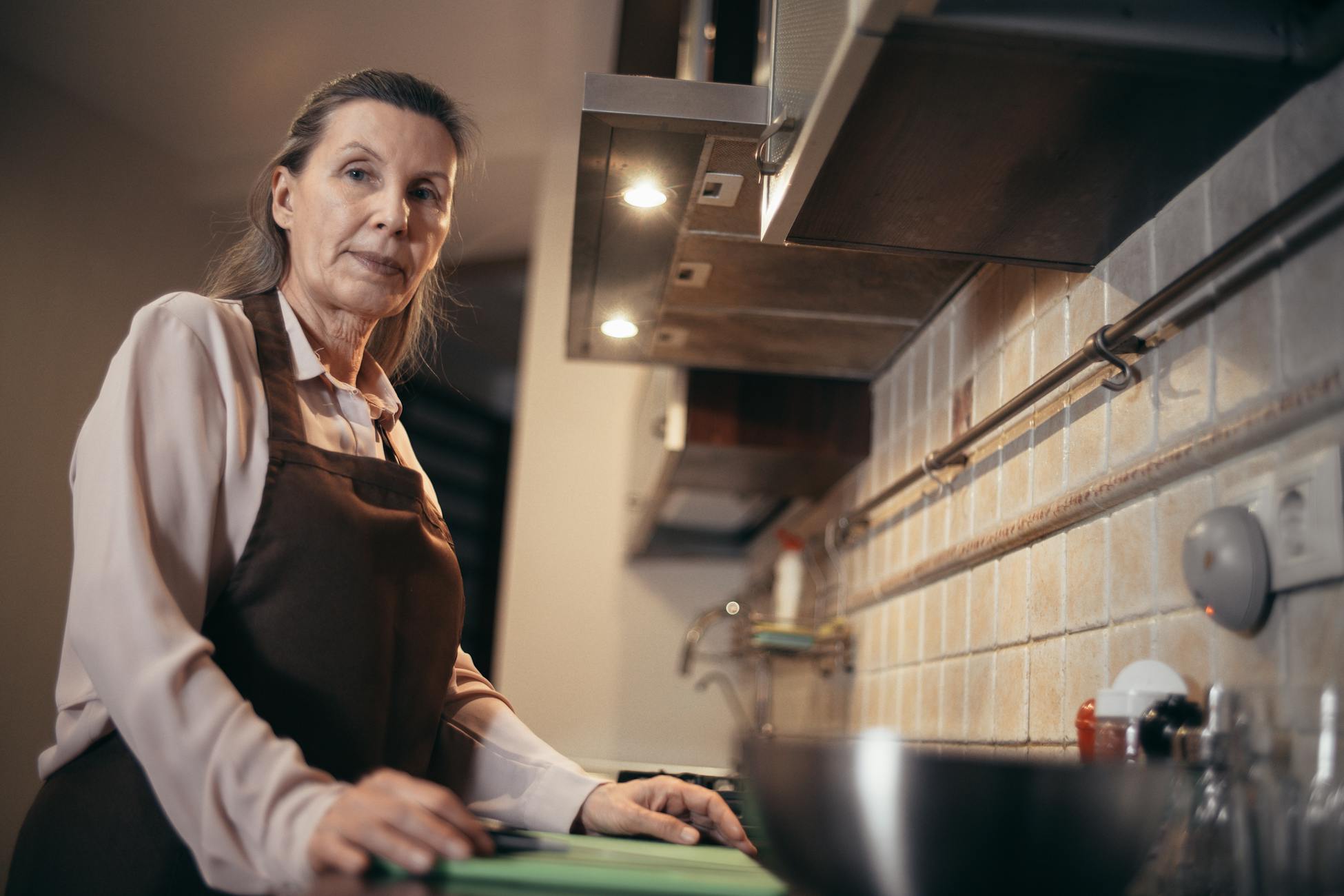 A mature woman in an apron stands confidently in a modern kitchen interior.
