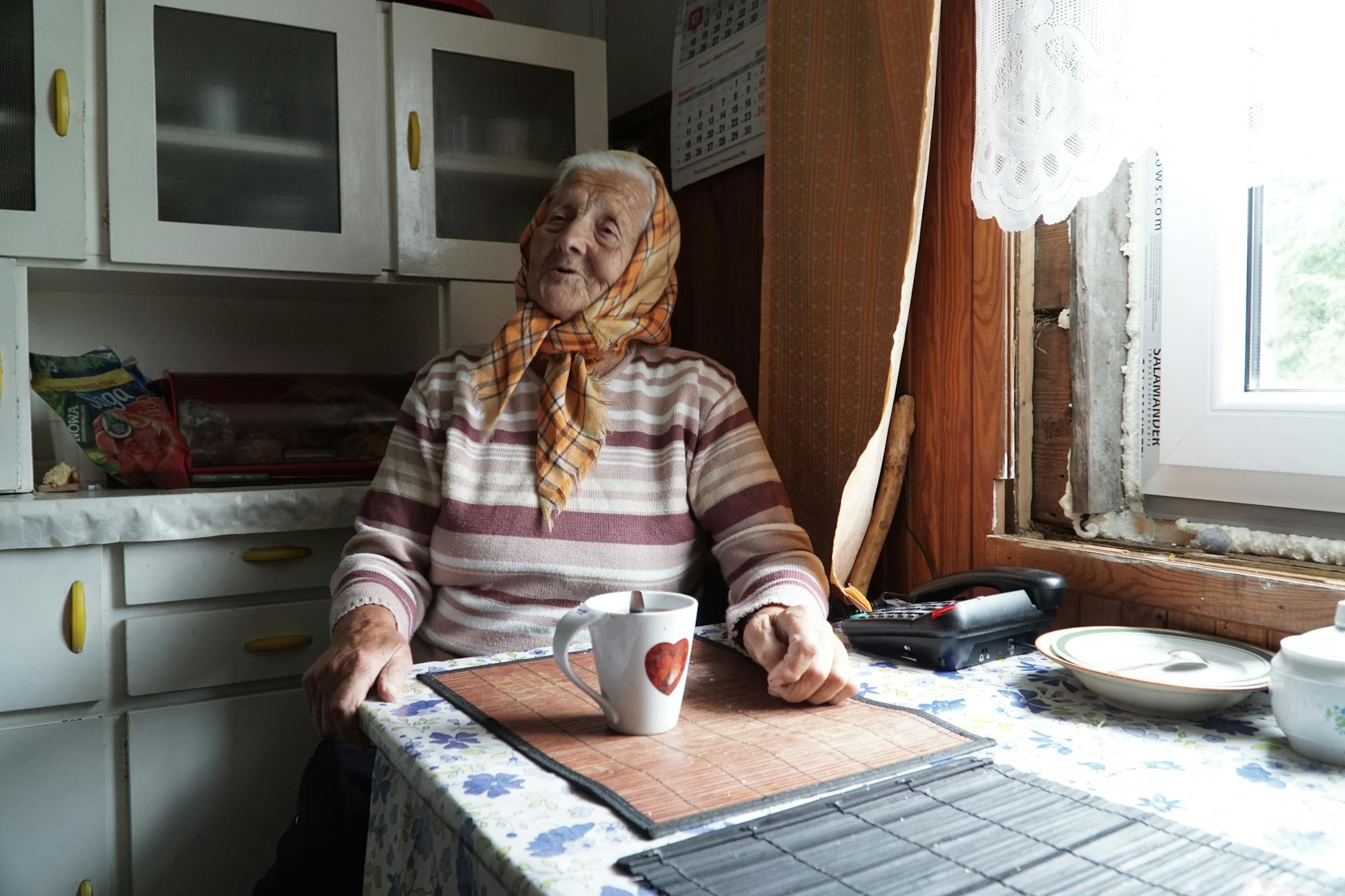 Elderly woman sitting at a table enjoying tea in a cozy kitchen with soft lighting.