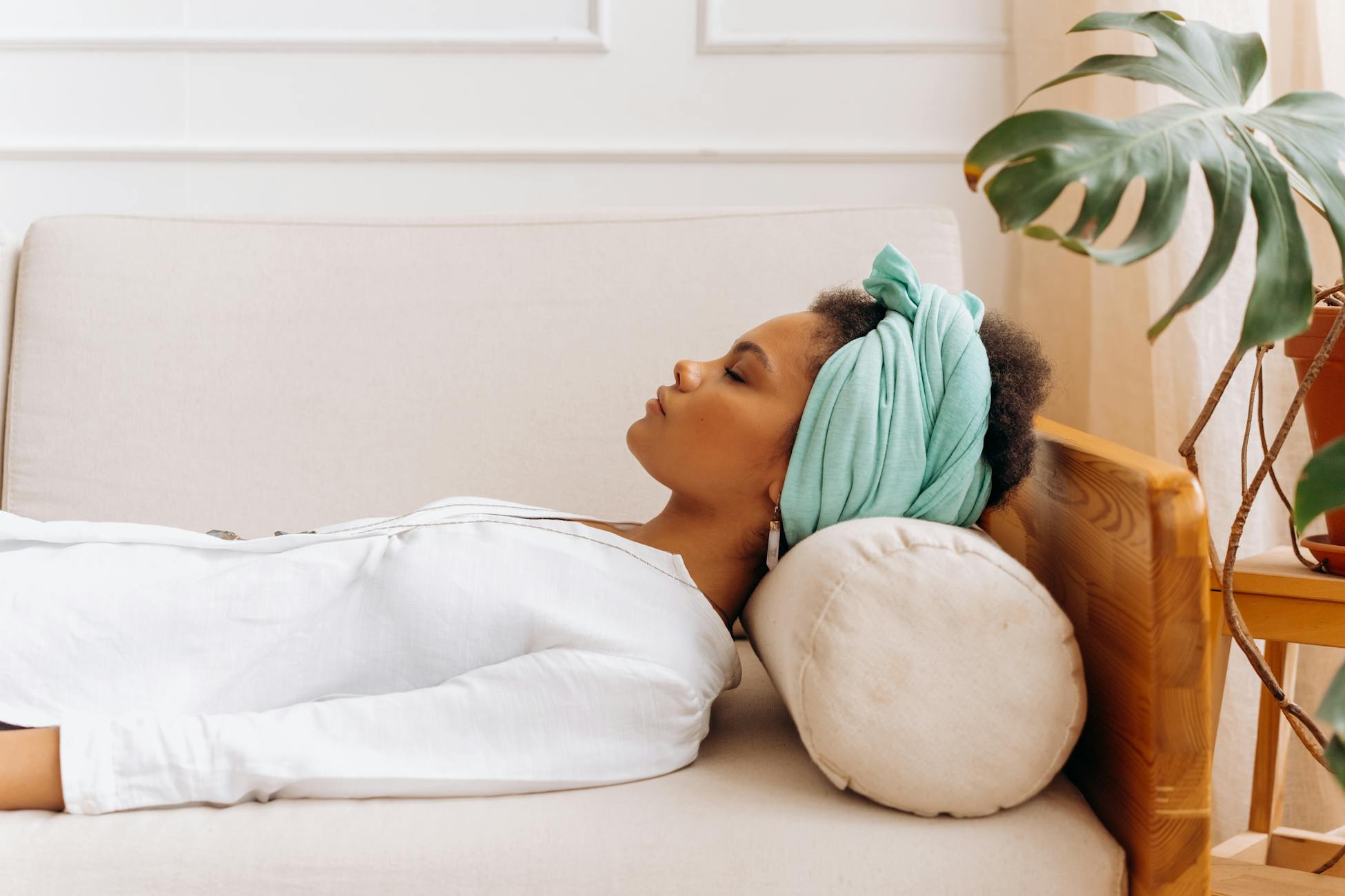A woman in a head scarf relaxes on a beige sofa indoors surrounded by plants, peaceful scene.