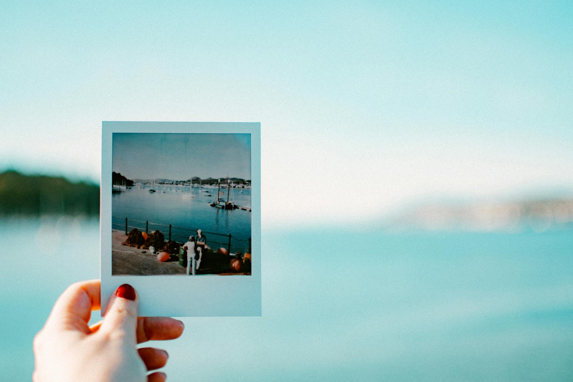 A hand holds a Polaroid photo of a serene waterfront view, with a background of calm waters and a clear sky.