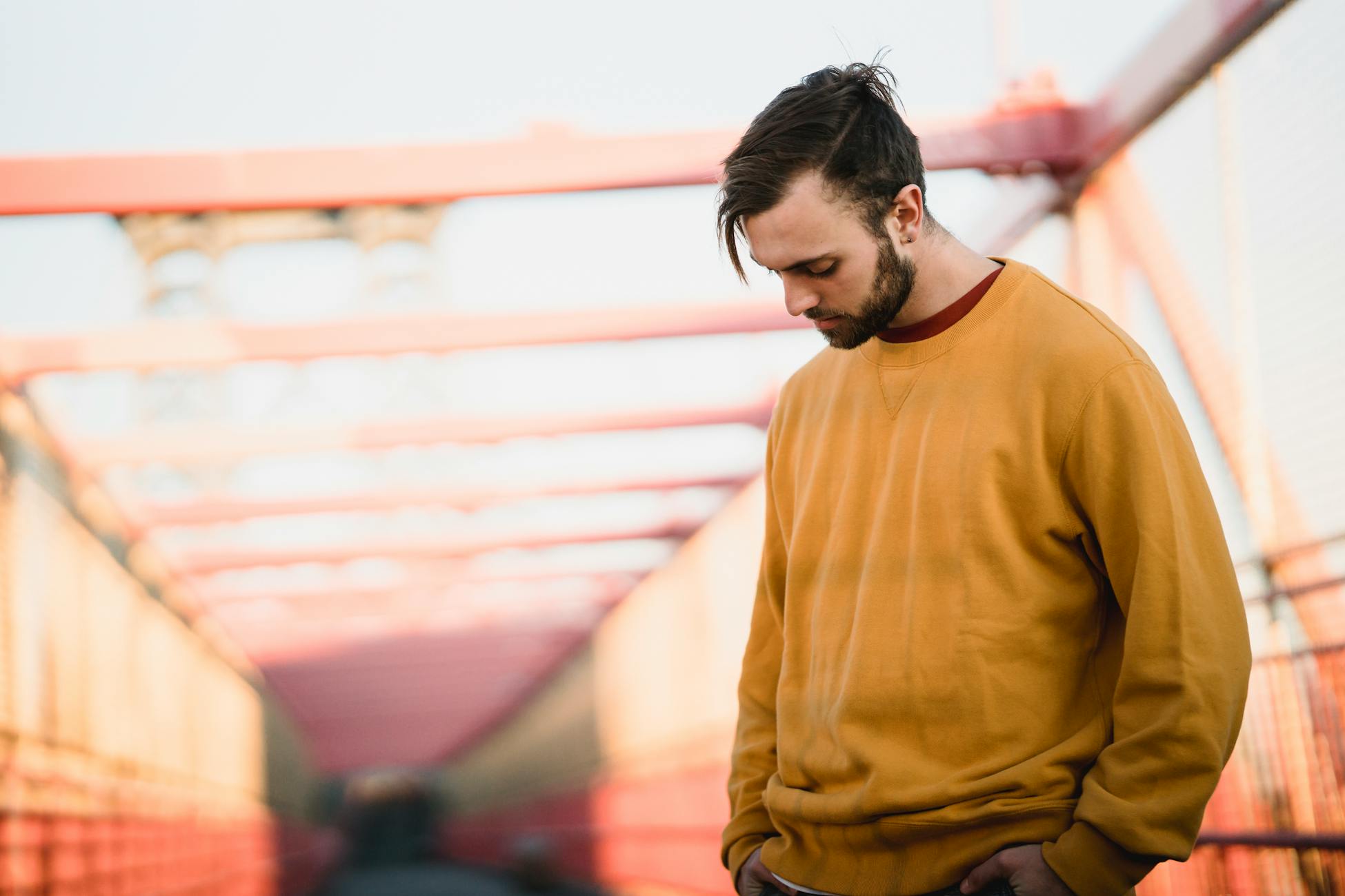 Young thoughtful bearded male in casual apparel with modern haircut looking down while standing with hands on hips in town