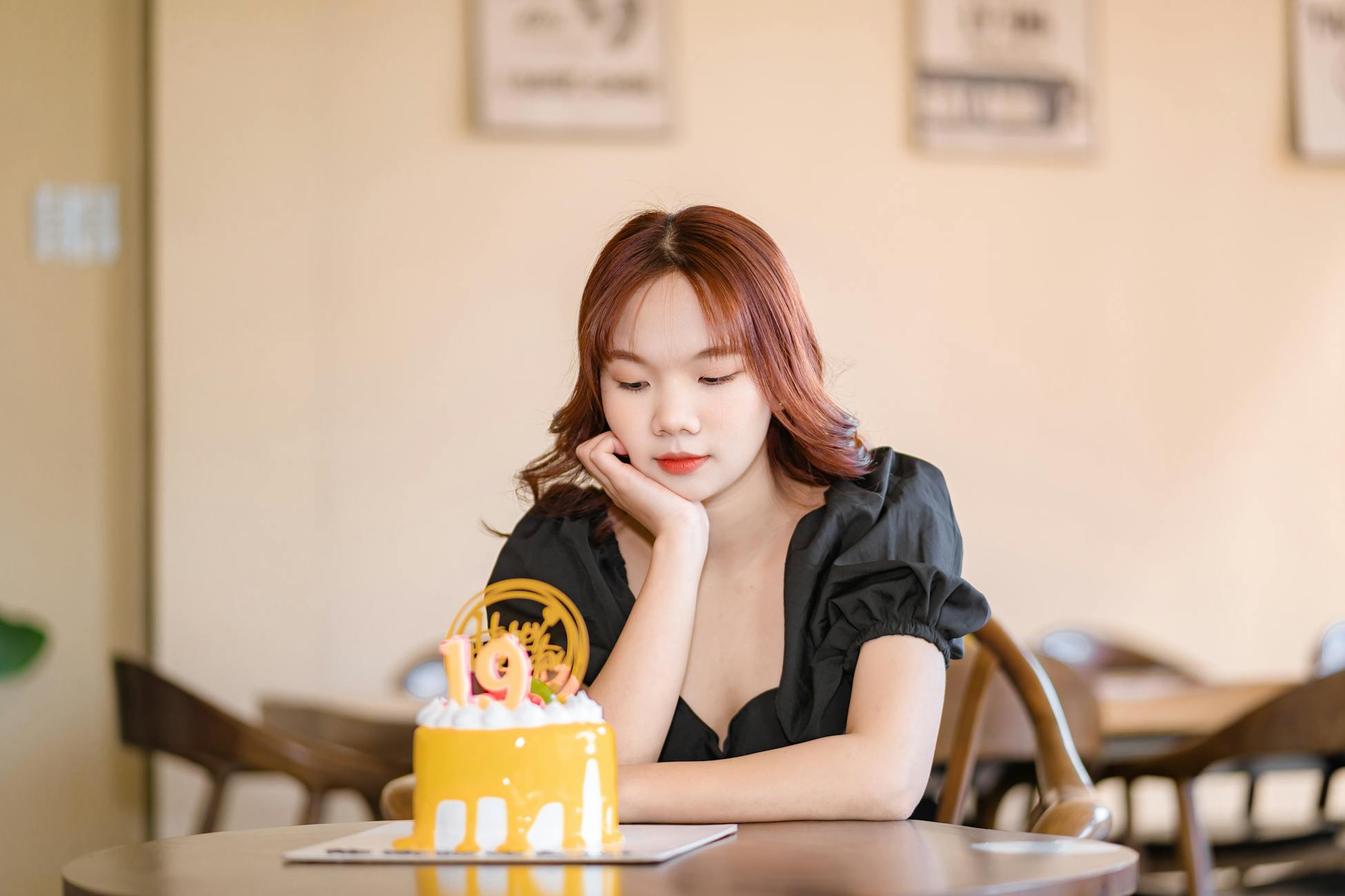 A thoughtful young woman sitting with a birthday cake featuring the number 19 in a café setting.