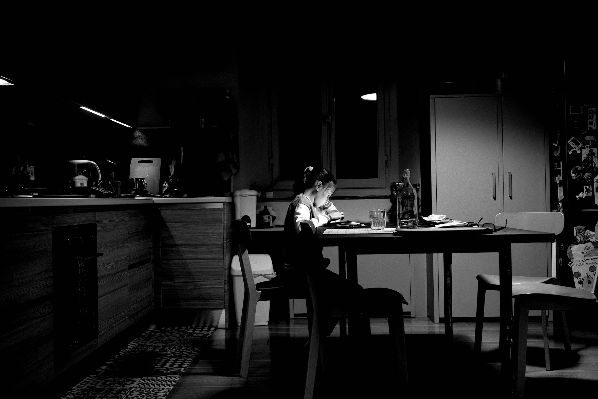 A child studying alone at a dimly lit kitchen table, creating a moody atmosphere.