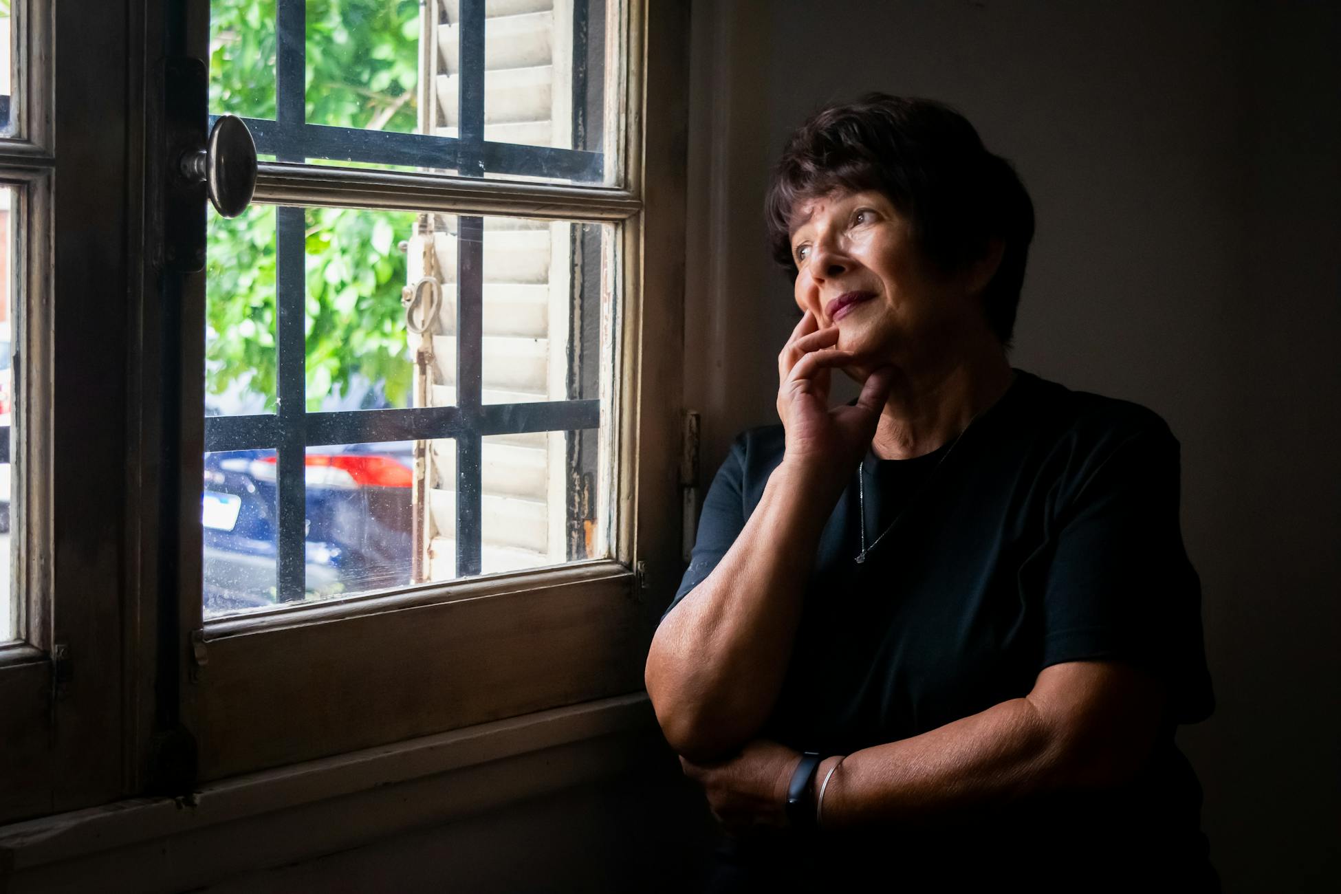 A thoughtful senior woman gazes out a window, reflecting softly indoors.