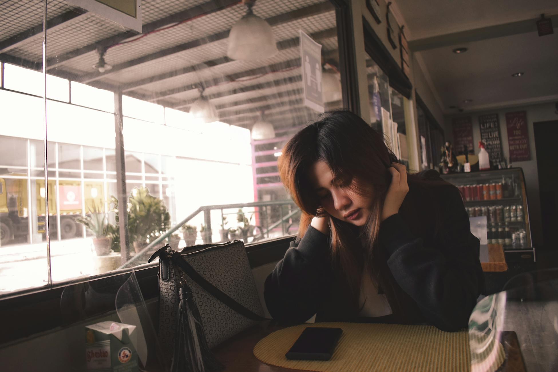 A woman deep in thought sits at a cafe table indoors with a phone beside her.