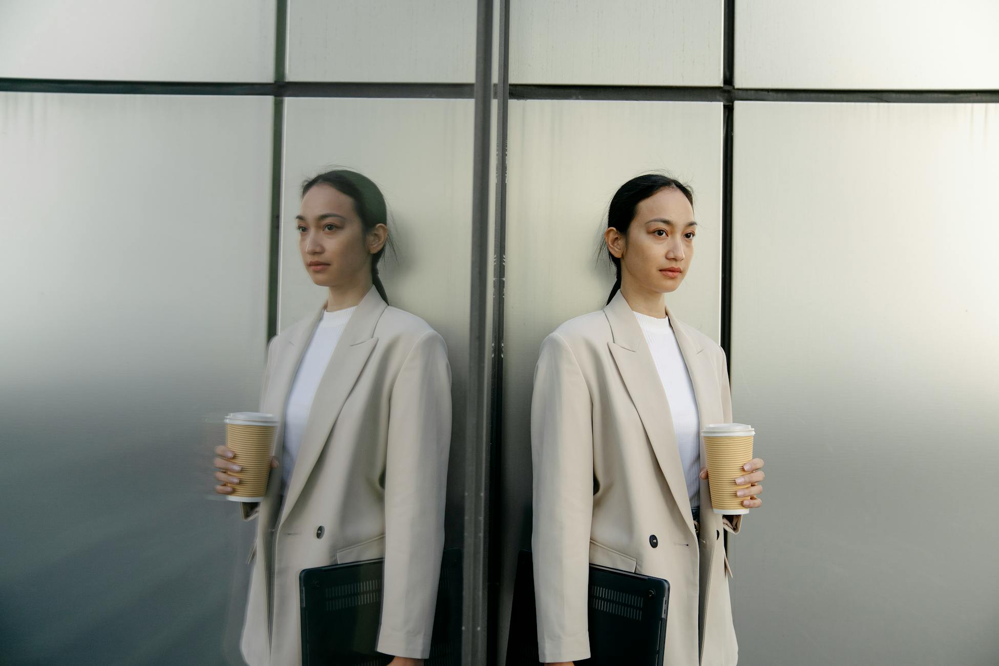 Emotionless Asian female office worker wearing formal clothes standing with paper cup of coffee and netbook near reflecting wall