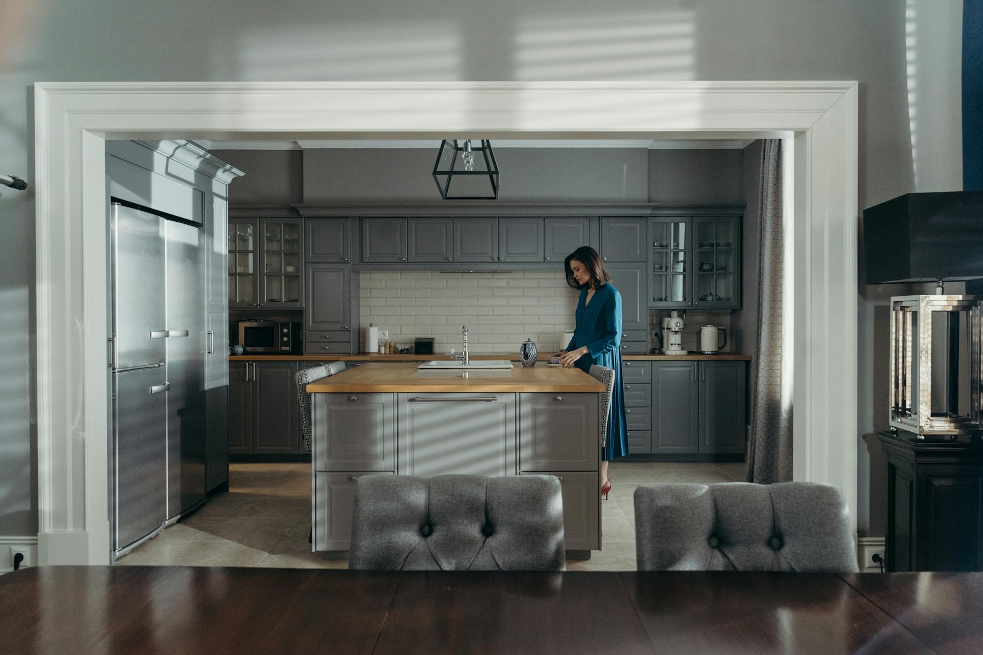 A woman in a blue dress stands in a modern, stylish kitchen, blending contemporary design with elegance.