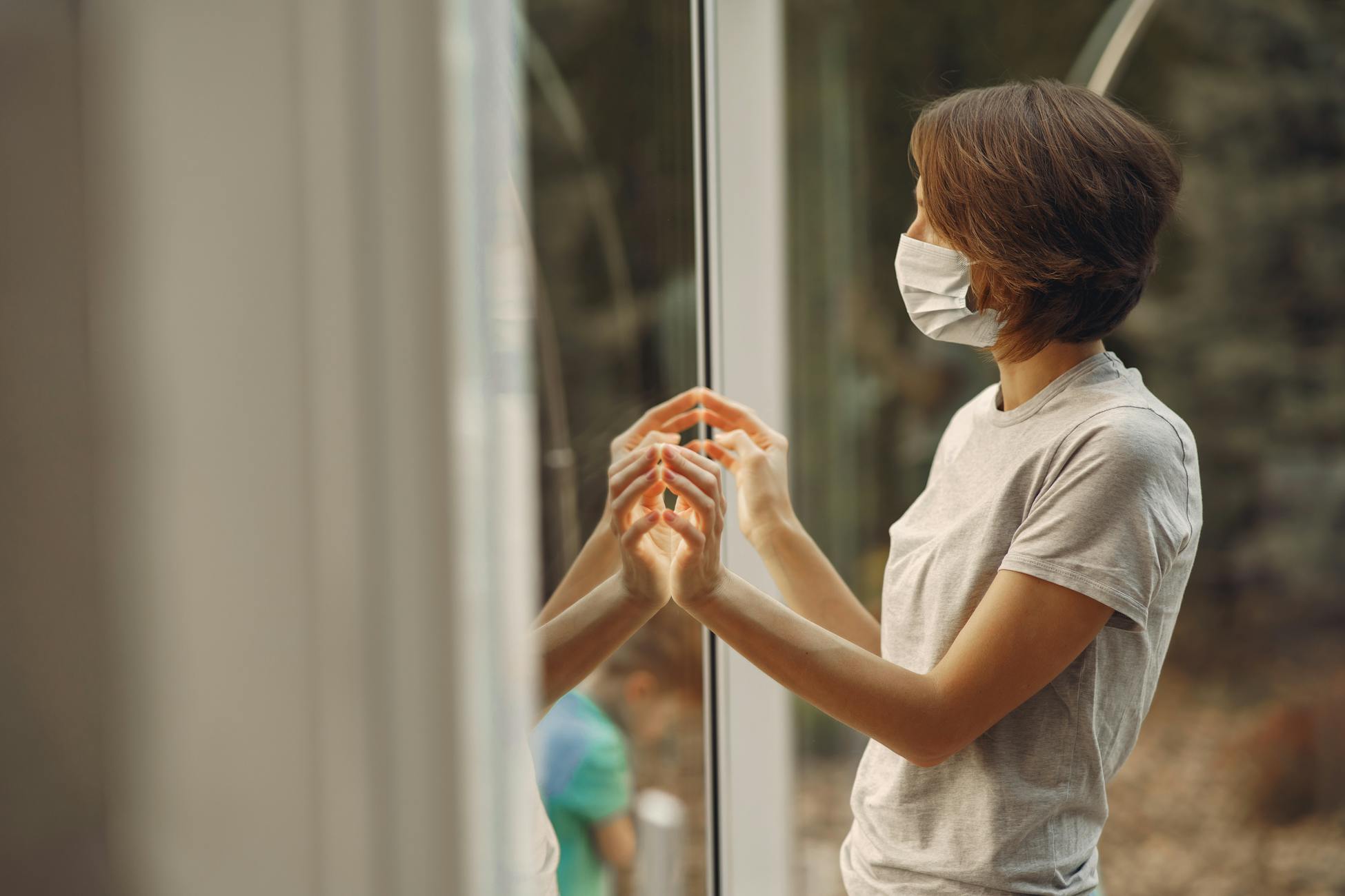 A woman in a face mask looks outside, her hands touching a glass window, reflecting in it.