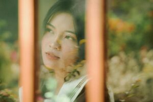 A thoughtful woman gazes outside through a glass window, surrounded by nature.