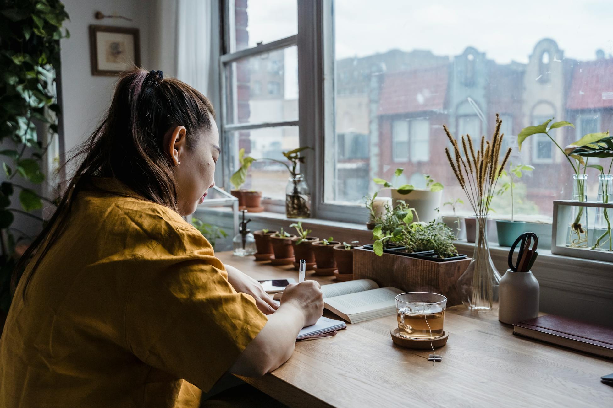 Asian woman writing in notebook in a cozy home office by window with plants.