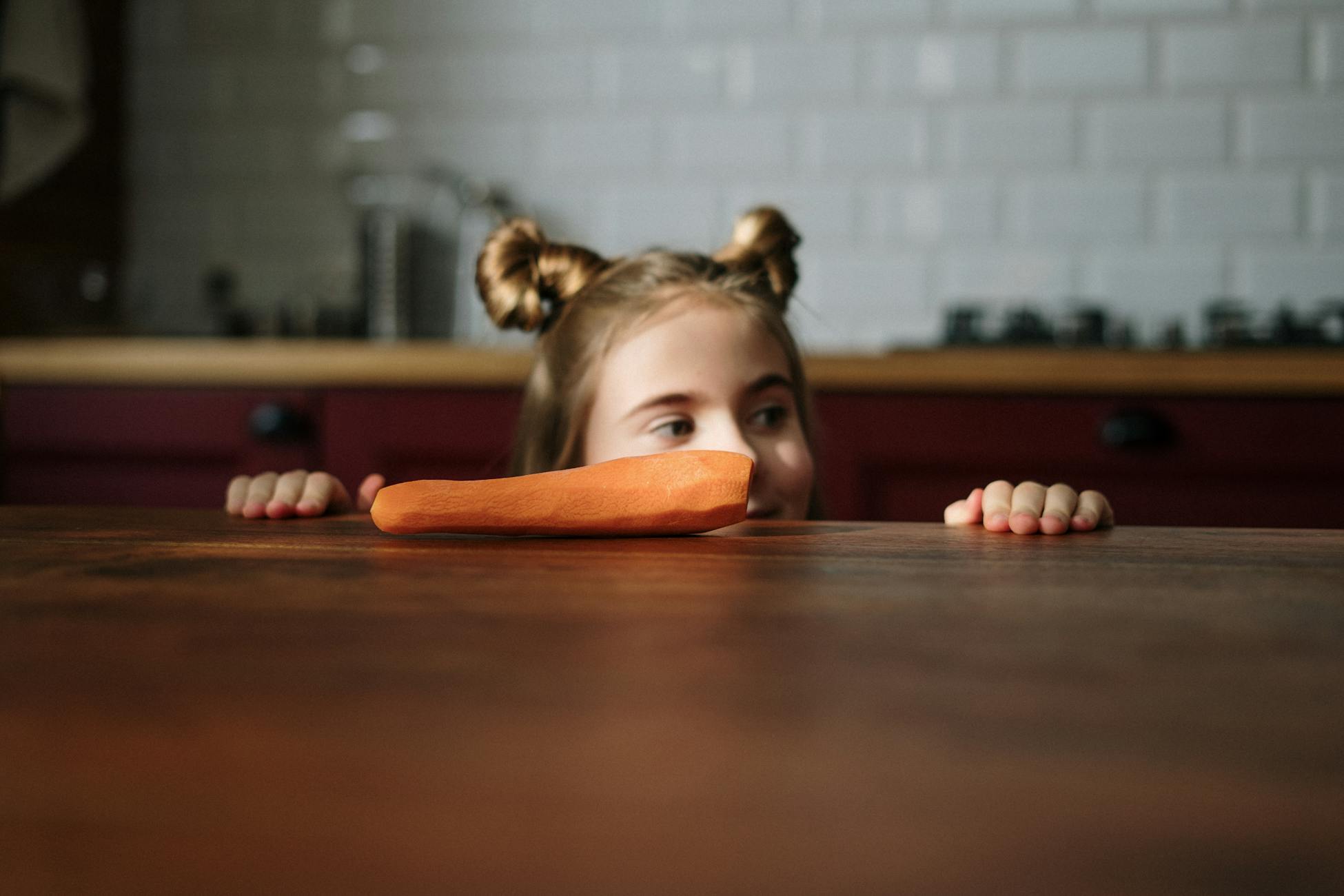 A curious child with a cute hairstyle peeks over a kitchen table at a fresh carrot.
