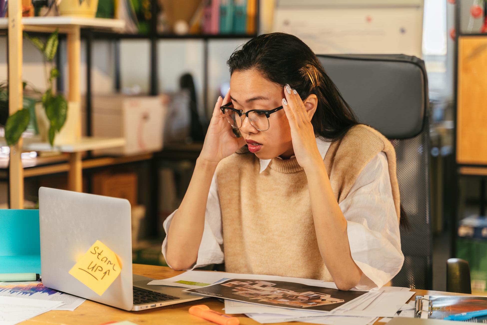 A woman with glasses shows stress while working at a laptop in an office environment.