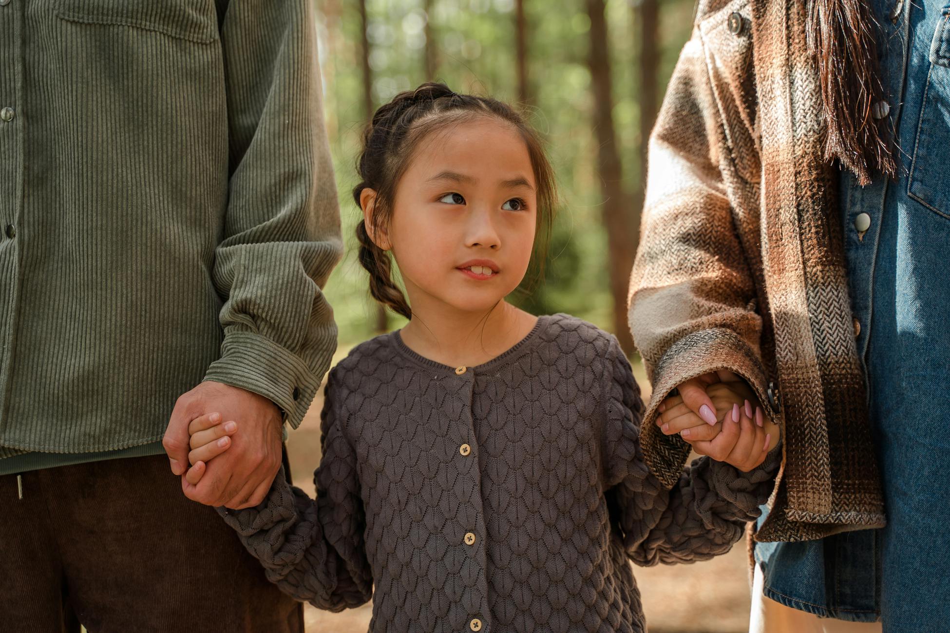 An adorable Asian girl holding hands with adults in a forest setting, expressing warmth and family love.