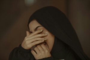 A young Muslim woman in black hijab smiling with her hands covering her face indoors.