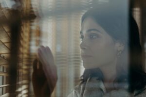 A thoughtful woman peeks through window blinds, creating a contemplative mood.