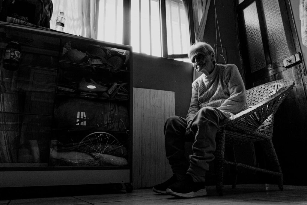 Black and white photo of a senior woman sitting alone in a dimly lit room, portraying solitude.