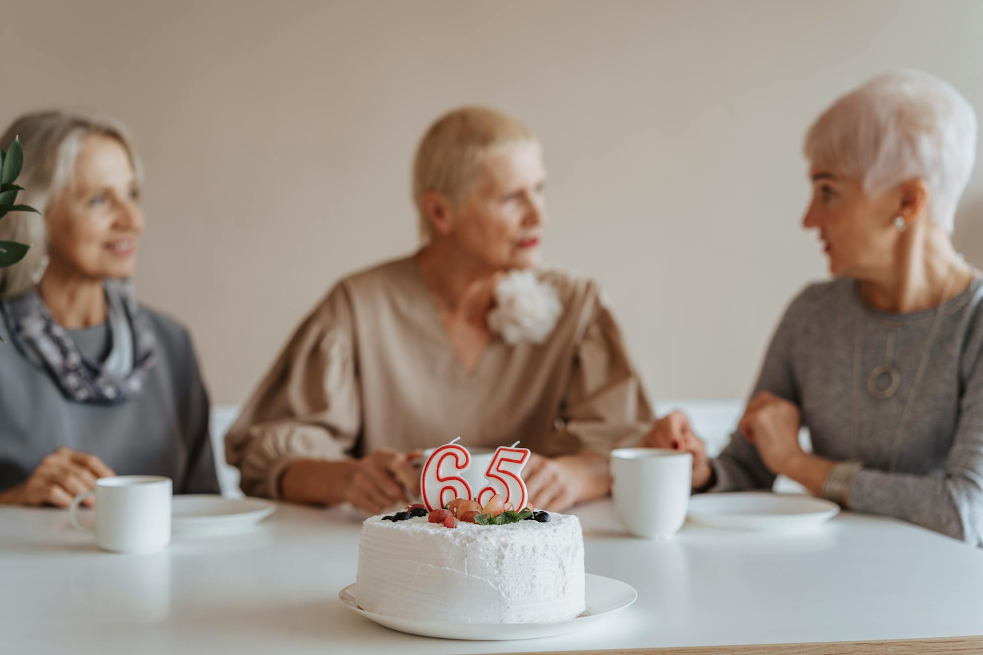 Three senior women celebrate a 65th birthday with cake and coffee indoors.