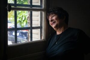 A senior woman looking thoughtfully out a window, framed by soft light and shadows.
