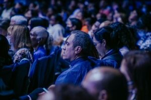 A large, diverse audience attentively listens during an indoor event.