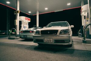 Two classic cars at a gas station at night, showcasing automotive design and urban ambiance.