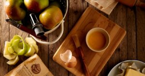 Apples, a pastry brush, glaze, and an old recipe book laid out on a wooden kitchen table