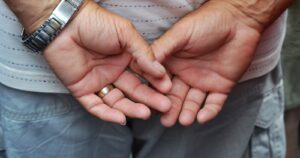 An older man's weathered hands clasped behind his back, wedding ring visible
