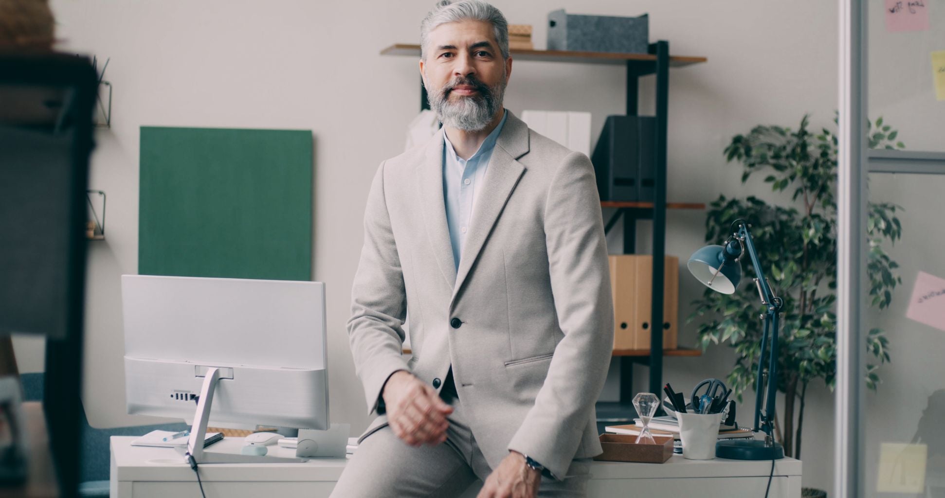A mature businessman in a beige suit sits confidently on a desk in a modern office setting