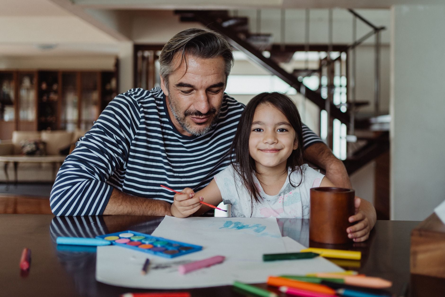Father and daughter sitting together, creating art with paints and pencils, fostering creativity and family bonding.